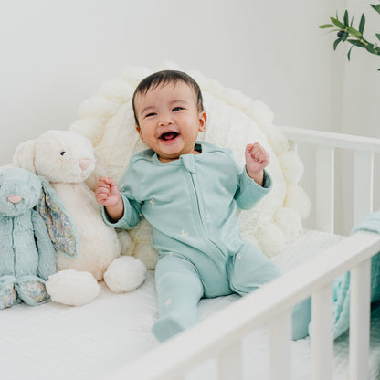 A happy baby wearing a mint blue organic zip sleepsuit in leaf print from Le Petit Society.