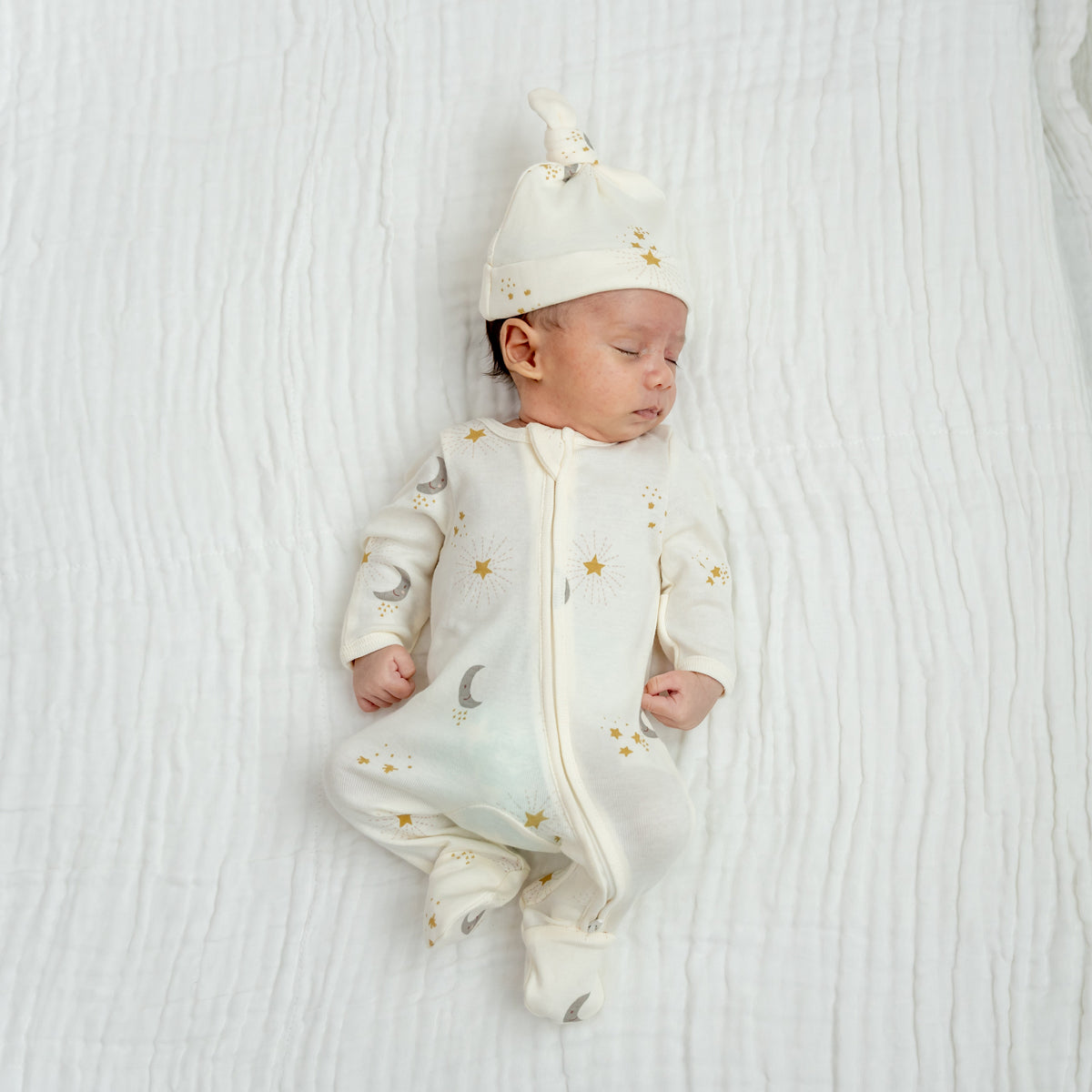 A newborn baby wearing an organic zip sleepsuit with foldable mittens and footies by Le Petit Society, featuring a print of moon and stars over a cream base.