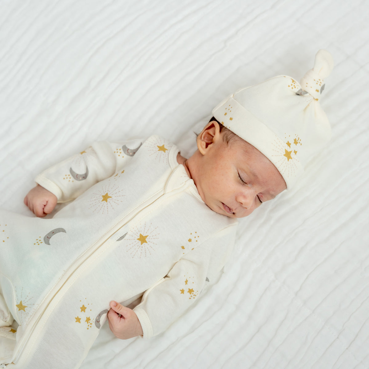 A newborn baby wearing an organic zip sleepsuit with foldable mittens and footies by Le Petit Society, featuring a print of moon and stars over a cream base.