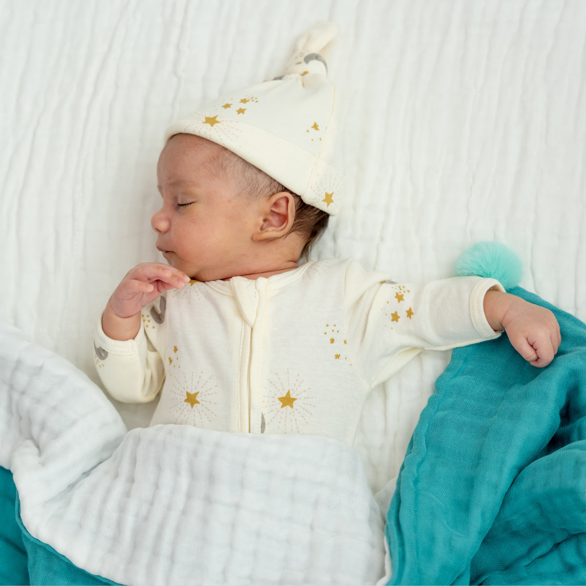 A newborn baby wearing an organic zip sleepsuit with foldable mittens and footies by Le Petit Society, featuring a print of moon and stars over a cream base.