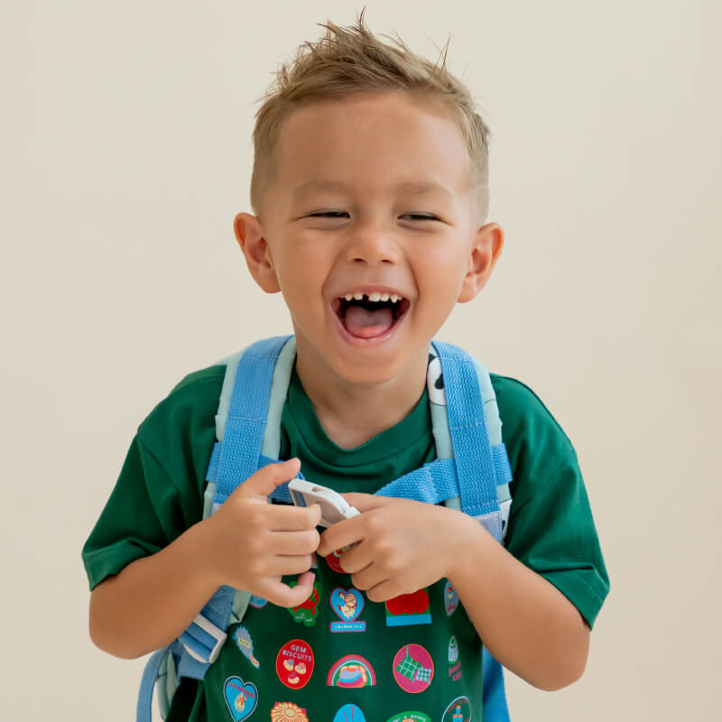 A laughing toddler boy holding on to the buckle clasp of his school bag.