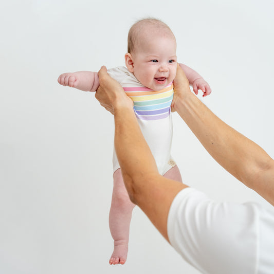 A baby in a short sleeved, white romper with a pastel rainbow print by Le Petit Society.