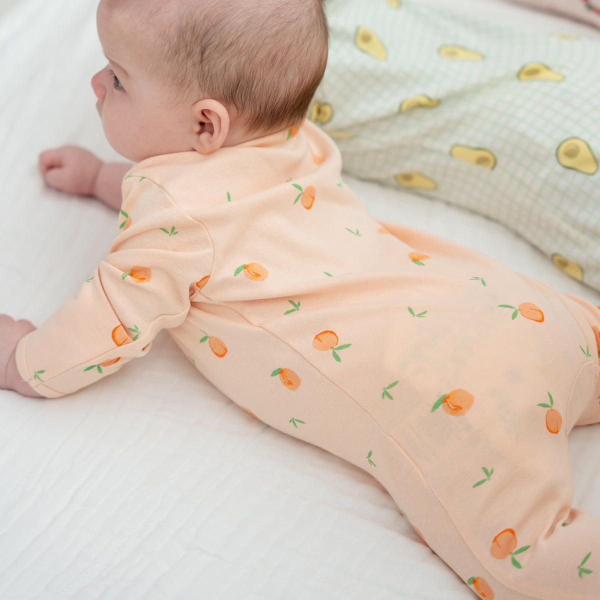 A baby in a coral pink zip sleepsuit with a print of peaches. 