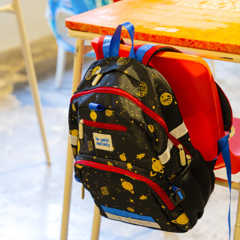 The kids single pocket school bag hanging on to a classroom chair with its red and blue shoulder straps.