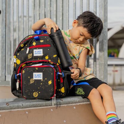 A primary school boy fitting his water bottle in the water bottle sleeve of the kids single pocket backpack in planet print.