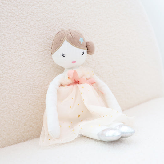 A plush doll of a ballerina in a low bun, pink dress, silver ballet shoes and blue pin sits against a sofa.