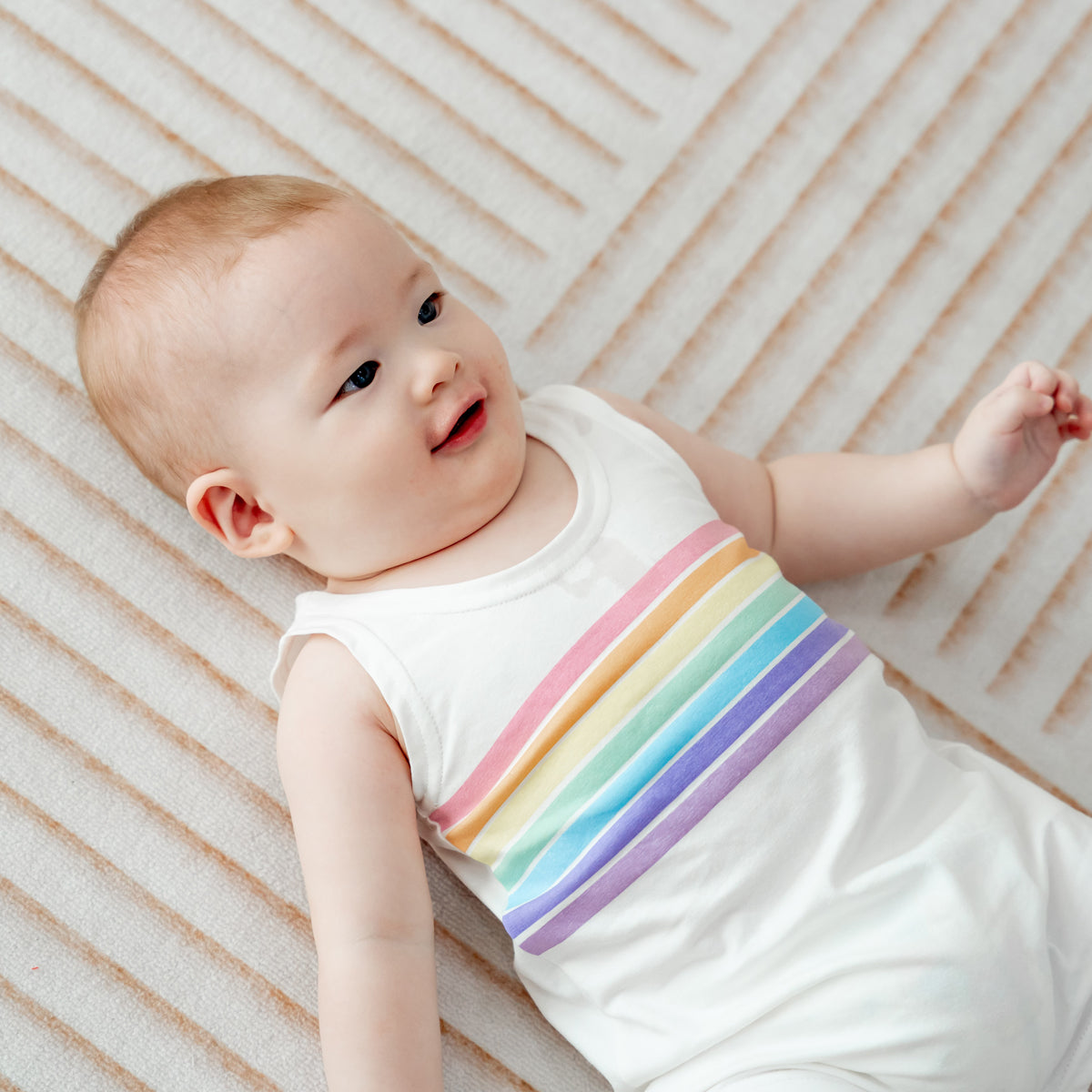 A baby boy in a sleeveless white romper with a pastel rainbow print by Le Petit Society.
