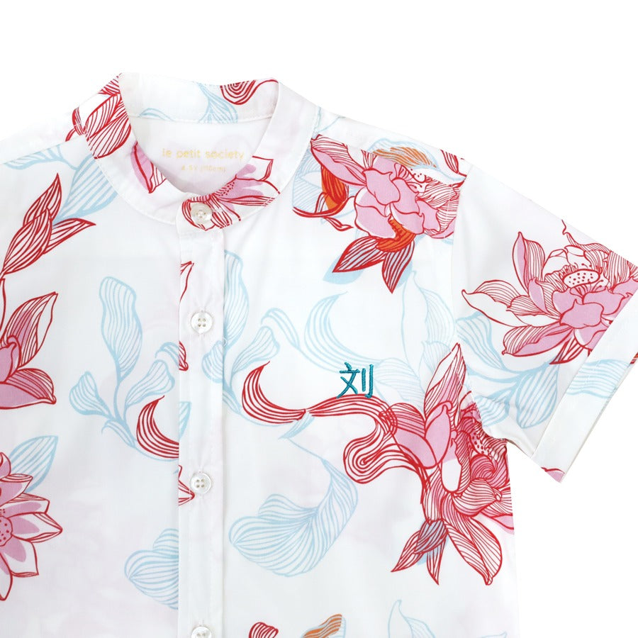 A close-up of the boys’ shirt from the Le Petit Society x Sheila Sim CNY 2026 Lotus Collection. The mandarin collar and button-down front are framed by hand-drawn lotus blossoms in coral and sky blue. A small teal embroidery detail is tucked beneath the p