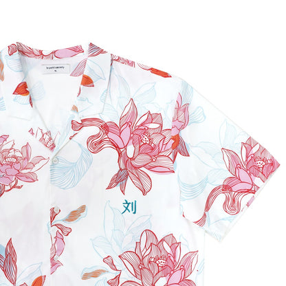 A close-up of the men’s shirt from the Le Petit Society x Sheila Sim CNY 2026 Lotus Collection. The airy white base showcases hand-drawn lotus blooms in layered coral, blush and sky-blue tones, arranged in sweeping, fluid strokes. The open collar and butt