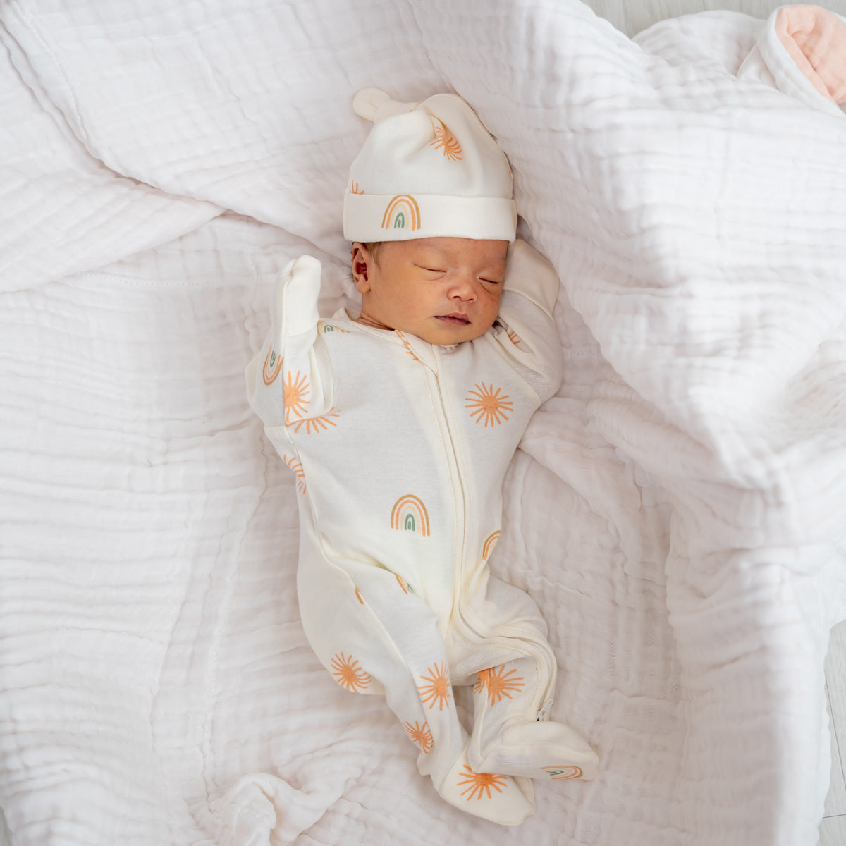 A newborn baby wearing an organic zip sleepsuit with foldable mittens and footies by Le Petit Society, featuring a print of sun and rainbows over a cream base. 