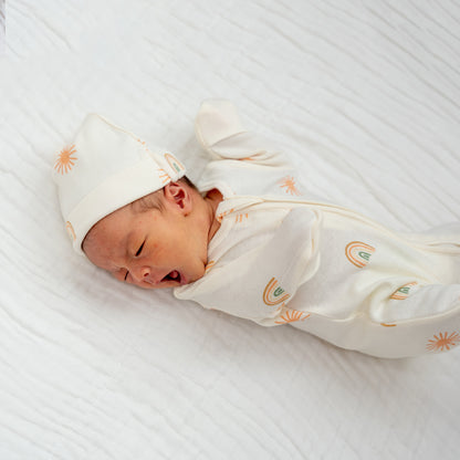 A newborn baby wearing an organic zip sleepsuit with foldable mittens and footies by Le Petit Society, featuring a print of sun and rainbows over a cream base. 