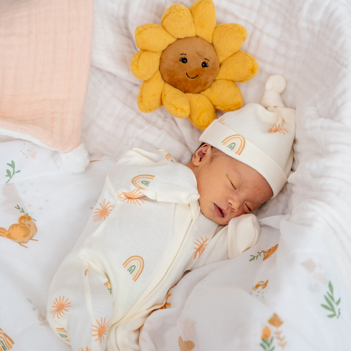A newborn baby wearing an organic zip sleepsuit with foldable mittens and footies by Le Petit Society, featuring a print of sun and rainbows over a cream base. 