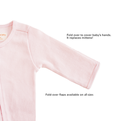 A baby organic zip sleepsuit in pink by Le Petit Society, with fold over flaps at the sleeves to cover a baby's hands, acting as a substitute for mittens.
