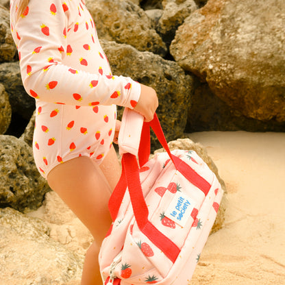 A little girl with a pink strawberry-printed swimsuit and thermal lunch bag from Le Petit Society.