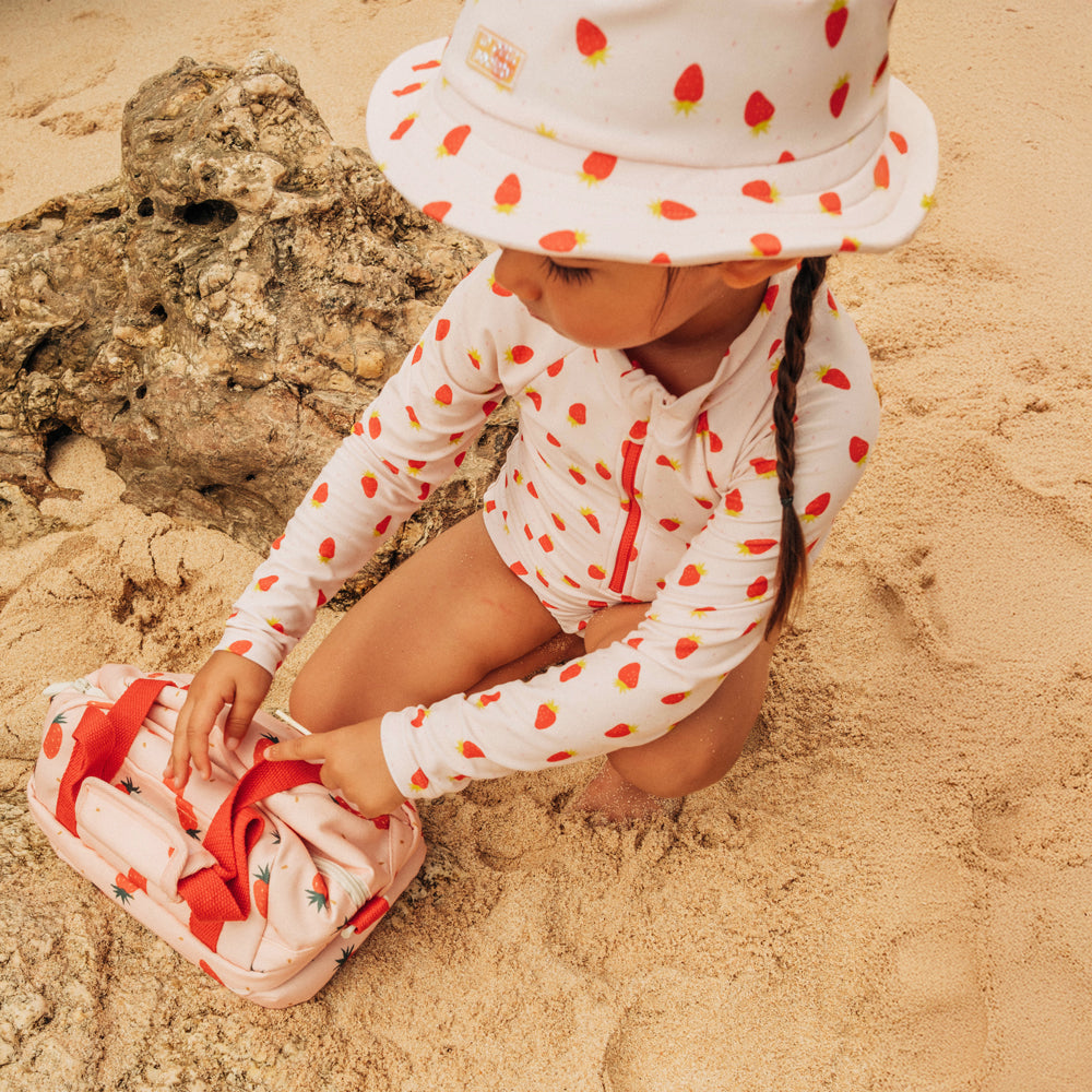 A little girl with a pink, strawberry-printed swimsuit and thermal lunch bag from Le Petit Society.