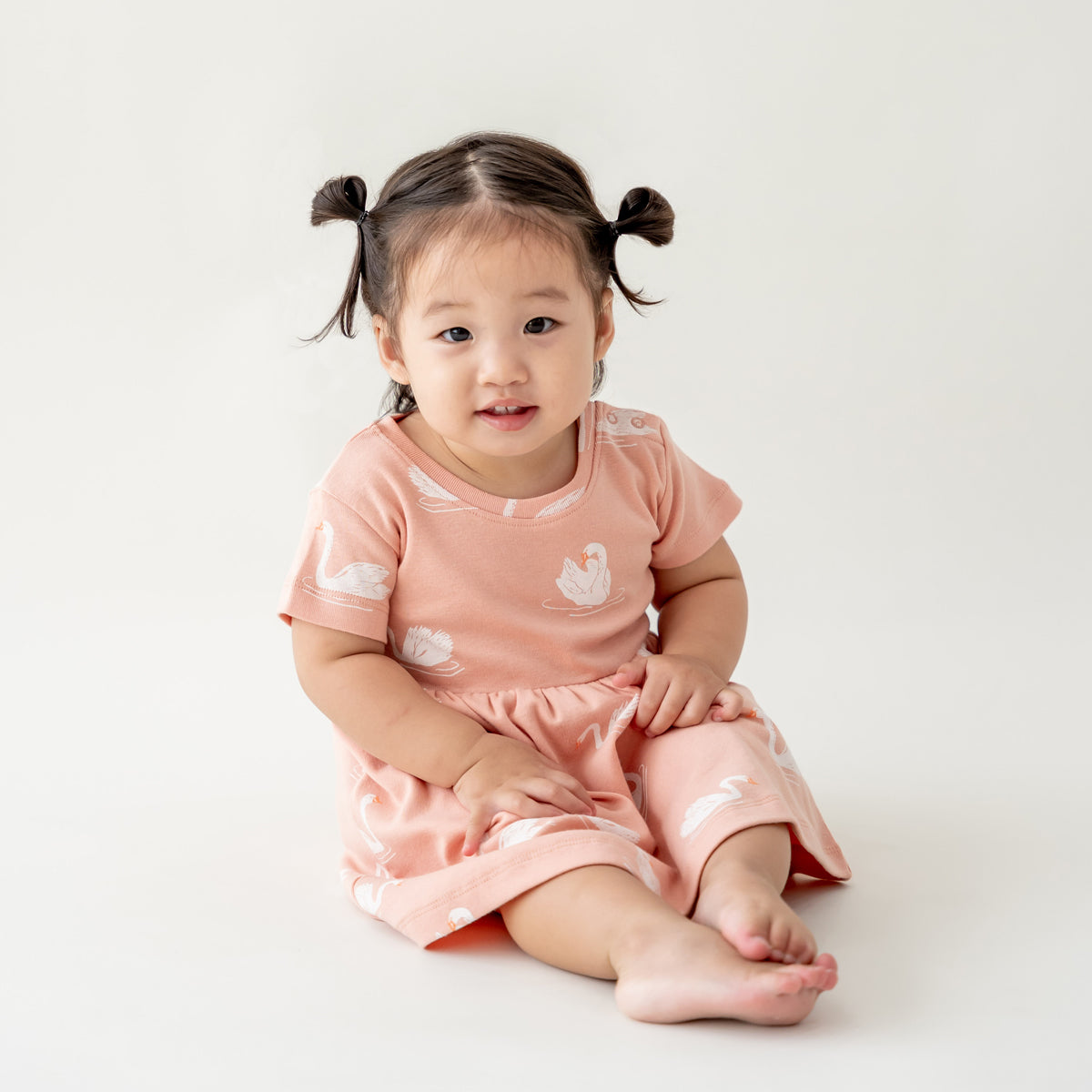 A girl toddler wearing a peach-coloured organic dress adorned with a print of swans from Le Petit Society.