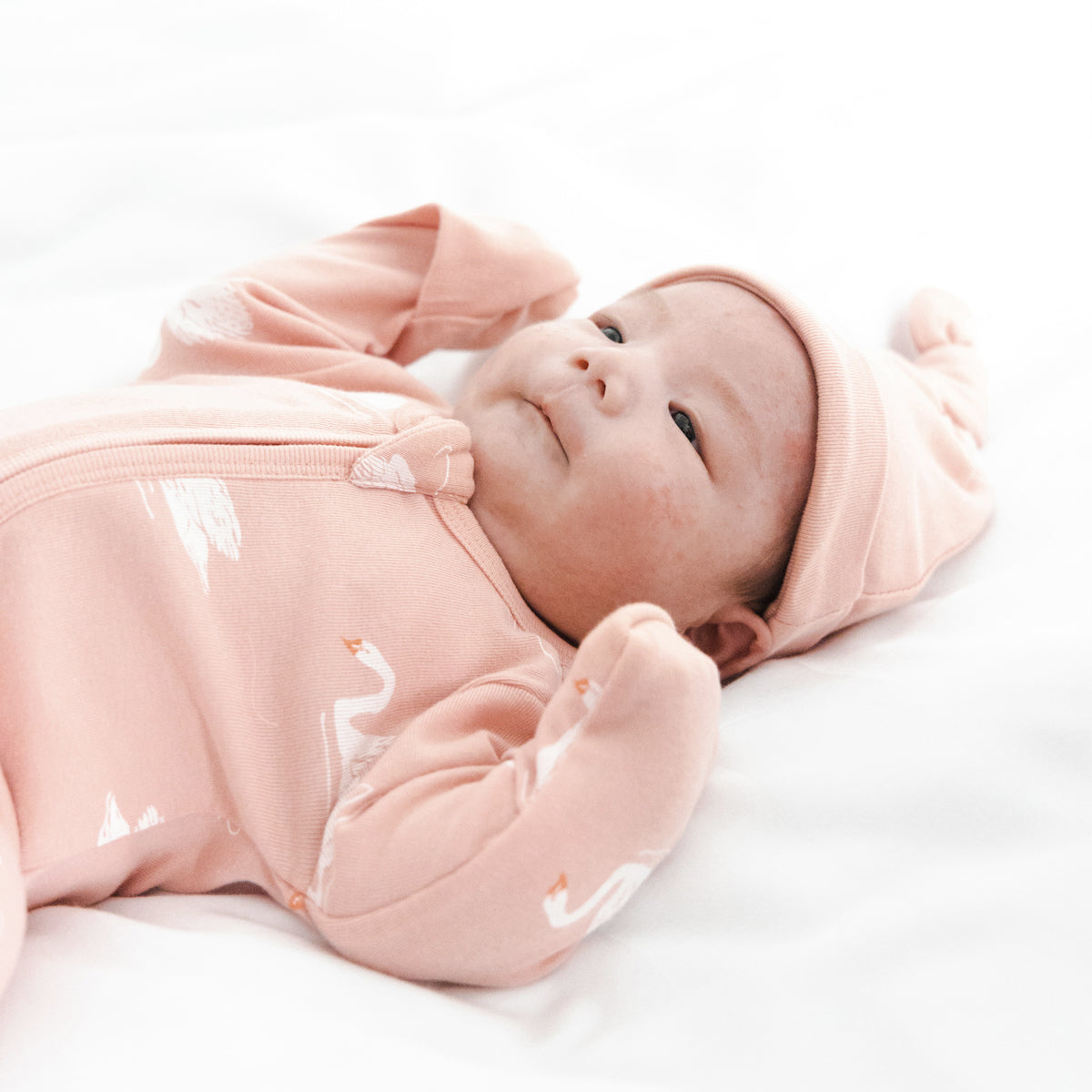 A newborn baby wearing an organic zip sleepsuit in peach with foldable mittens and footies by Le Petit Society, featuring a print of swans.