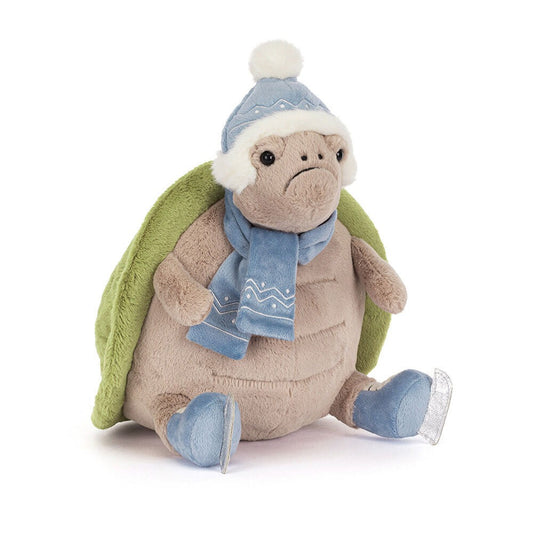 A 45 degree view of a plush turtle toy with a green shell, wearing a blue winter hat with a white pom-pom, a matching blue scarf, and blue skates. The turtle has a soft, beige body and a cute, slightly grumpy expression