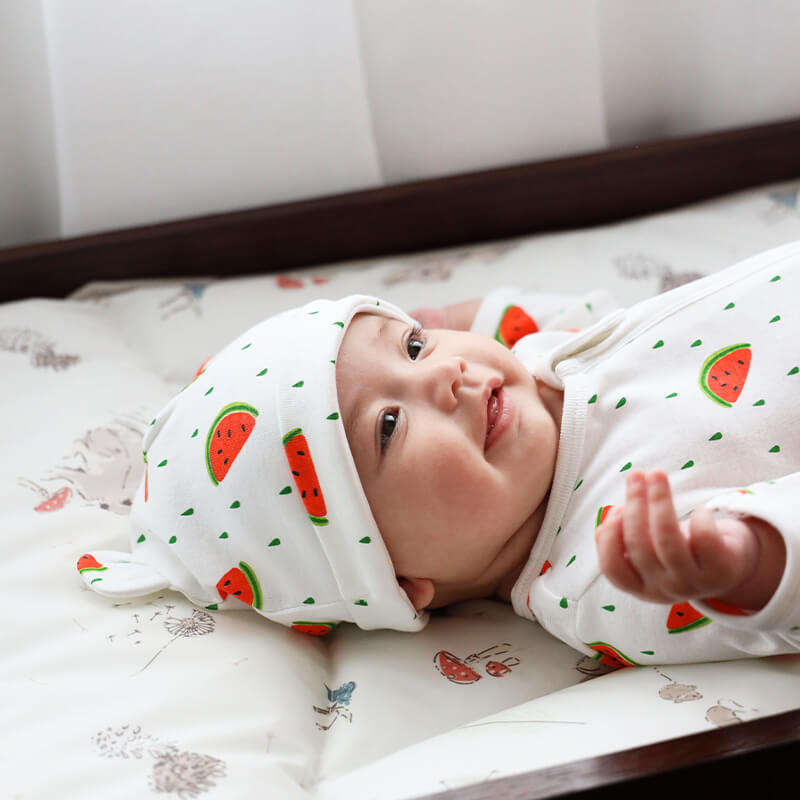 A baby wearing Le Petit Society's organic zip sleepsuit and bear hat in watermelon print.